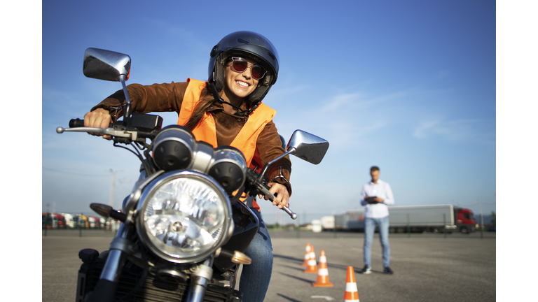 Female student with helmet taking motorcycle lessons and practicing ride. In background traffic cones and instructor with checklist rating and evaluating the ride. Motorcycle school of driving.