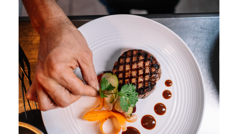 Hand of a chef garnishing a fillet of beef with herbs and vegetables on a ceramic plate in a kitchen