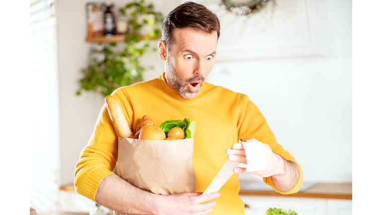 Surprised man looking at store receipt after shopping, holding a paper bag with healthy food. Guy in the kitchen.