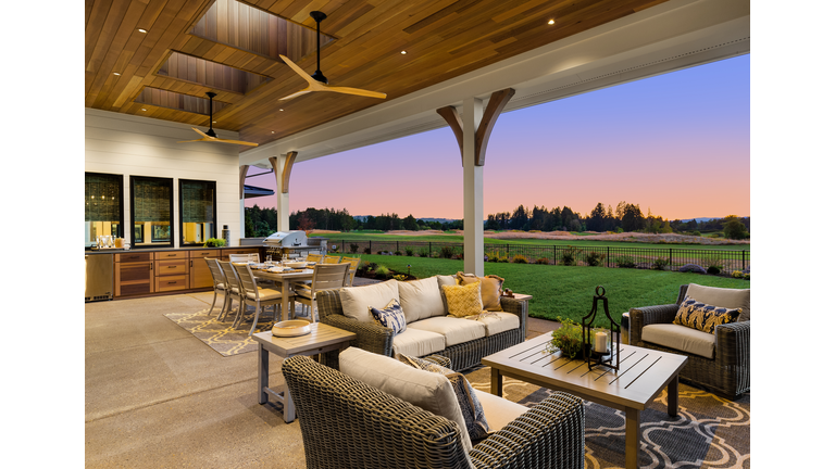 Luxury home exterior at sunset: Outdoor covered patio with kitchen, barbecue, dining table, and seating area, overlooking grass field and trees.