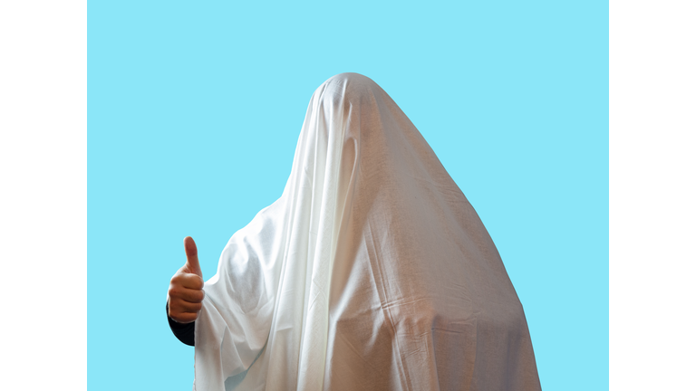 A man covered with a white sheet like a ghost shows a thumb on a blue background