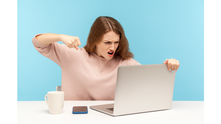 Enraged furious woman in casual clothes roaring madly and punching laptop screen, threatening with fist