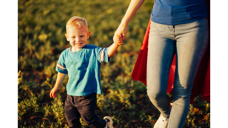 Super mom and her son walk forward holding hands. Cheerful family, a woman in a red raincoat as a superhero