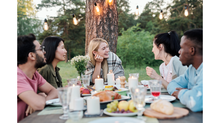 Young joyful couples eating and talking by served festive table under pine tree