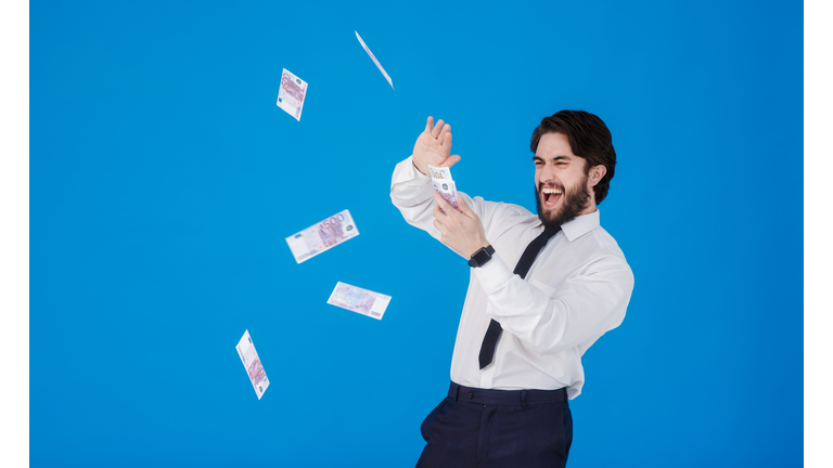 A young cheerful bearded businessman in a white shirt and black tie on a blue background splashes money. Crazy guy throws bills of money. Studio isolated portrait