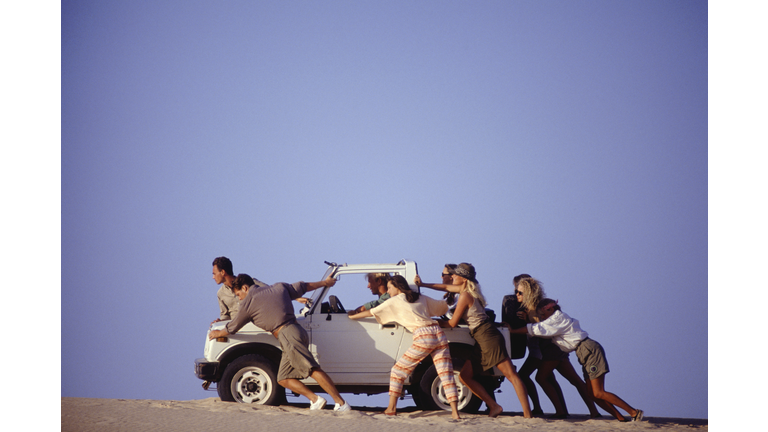 Group of young people pushing broken down car across sand, side view