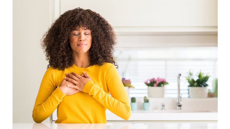 African american woman wearing yellow sweater at kitchen smiling with hands on chest with closed eyes and grateful gesture on face. Health concept.