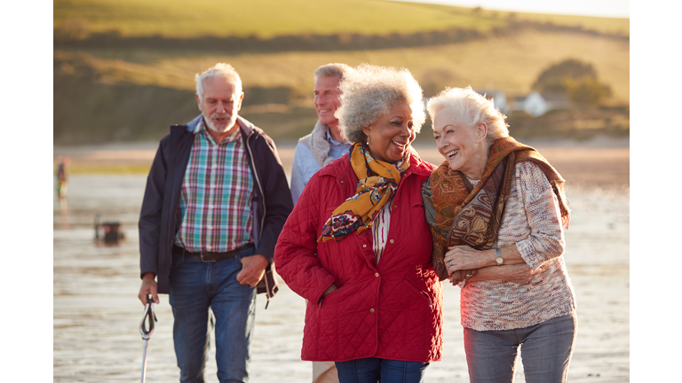 Group Of Smiling Senior Friends Walking Arm In Arm Along Shoreline Of Winter Beach