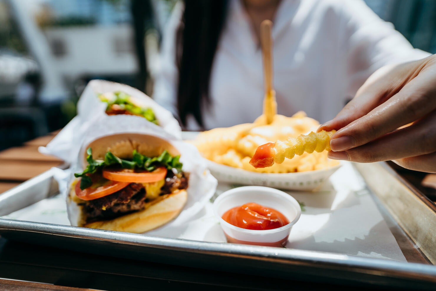 Close up of young Asian woman dipping fries with ketchup and eating beef burger in an outdoor fast food restaurant