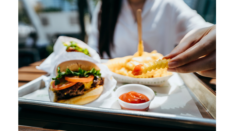 Close up of young Asian woman dipping fries with ketchup and eating beef burger in an outdoor fast food restaurant