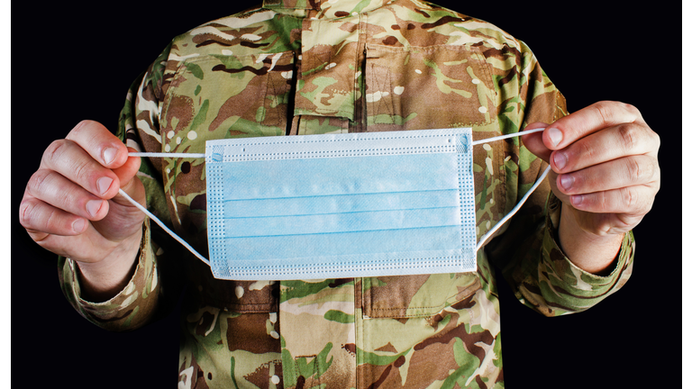 Soldier in camouflaged uniform holding a medical protective face mask on black background.