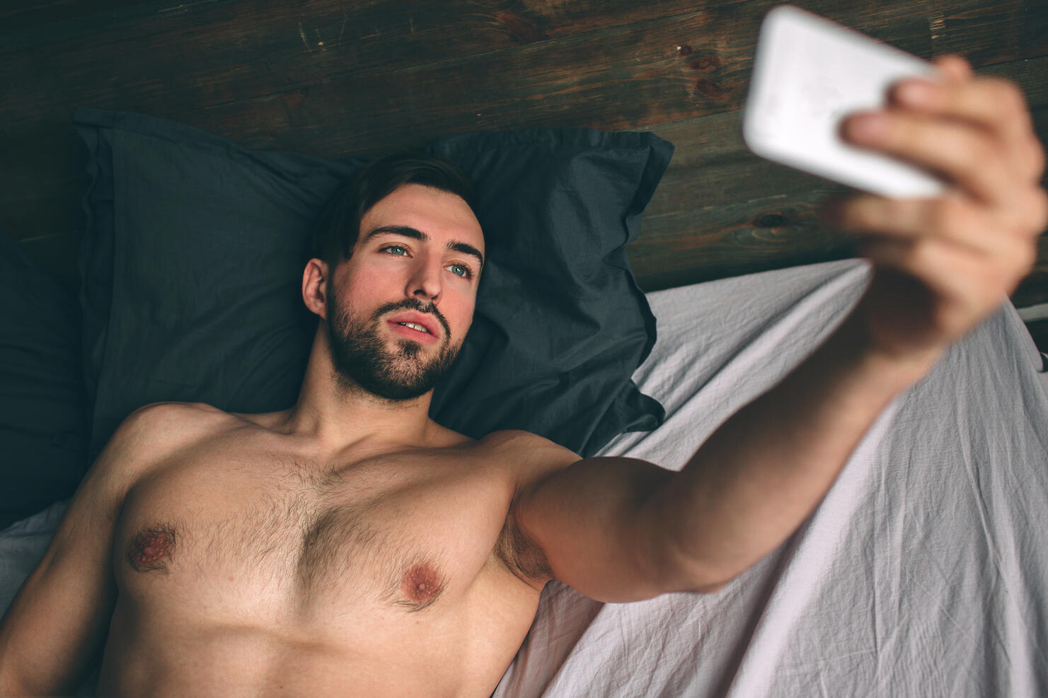 Naked bearded dark-hair handsome man shirtless in white bed taking a selfie