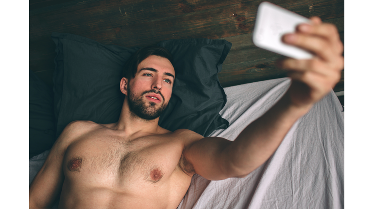 Naked bearded dark-hair handsome man shirtless in white bed taking a selfie