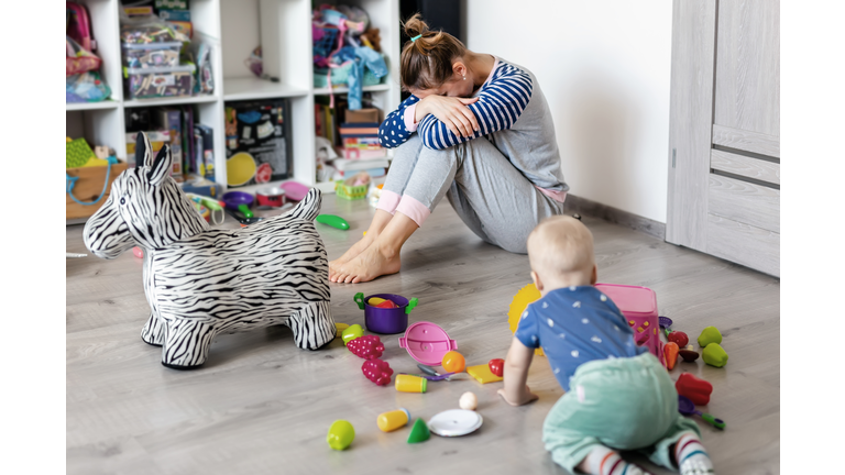 Tired of everyday household mother sitting on floor with hands on face. Kid playing in messy room. Scaterred toys and disorder. Happy parenting