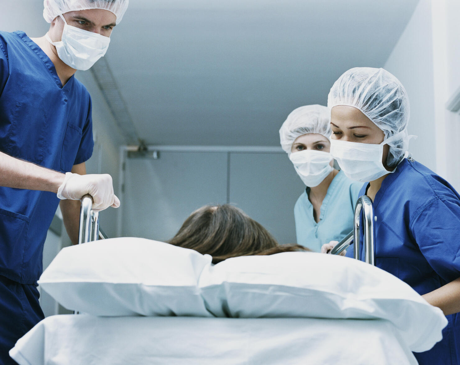 Three Surgeons in Scrubs and Protective Masks Look Down at a Patient on a Hospital Trolley
