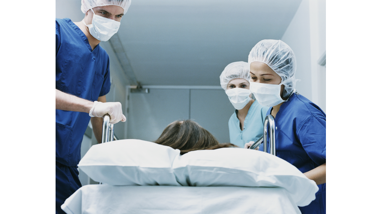 Three Surgeons in Scrubs and Protective Masks Look Down at a Patient on a Hospital Trolley