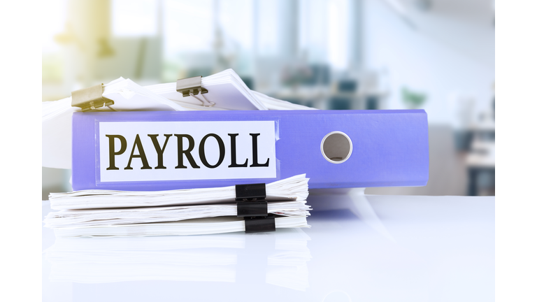 Payroll, the term is written on a folder of documents in trendy purple, lying on a stack of documents on an office table against the backdrop of an office with a soft blurred background.