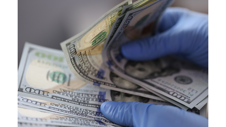 Hands in blue gloves hold one hundred dollar bills