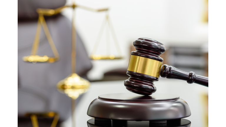 Wooden judge gavel or a wood hammer and a soundboard used by a judge person on a desk in a courtroom with a blurred brass scale of justice behind.