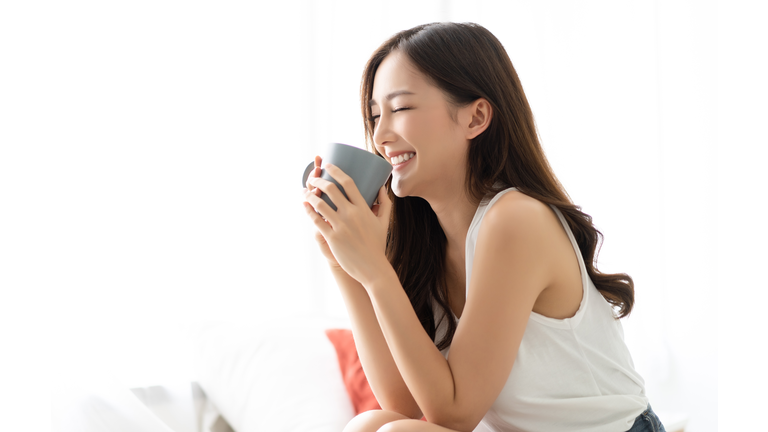 Happy smiling beautiful Asian woman awaking with cup of coffee at bedroom with Eyes Closed. People Lifestyle concept;