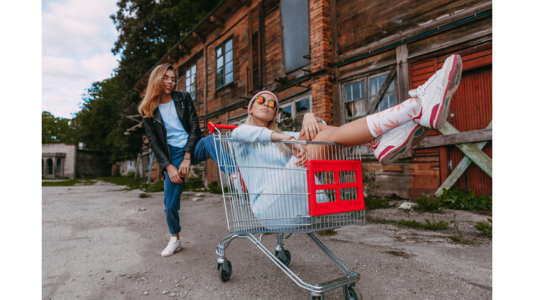 A long haired woman wearing a powder blue dress, pink woollen hat is chilling with her female friend, who is sitting on a shopping trolley, wearing a black leather jacket and blue jeans, in a derelict trading estate