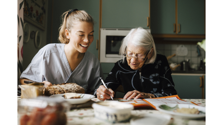 Senior woman solving crossword puzzle in book sitting by smiling nurse in kitchen at home