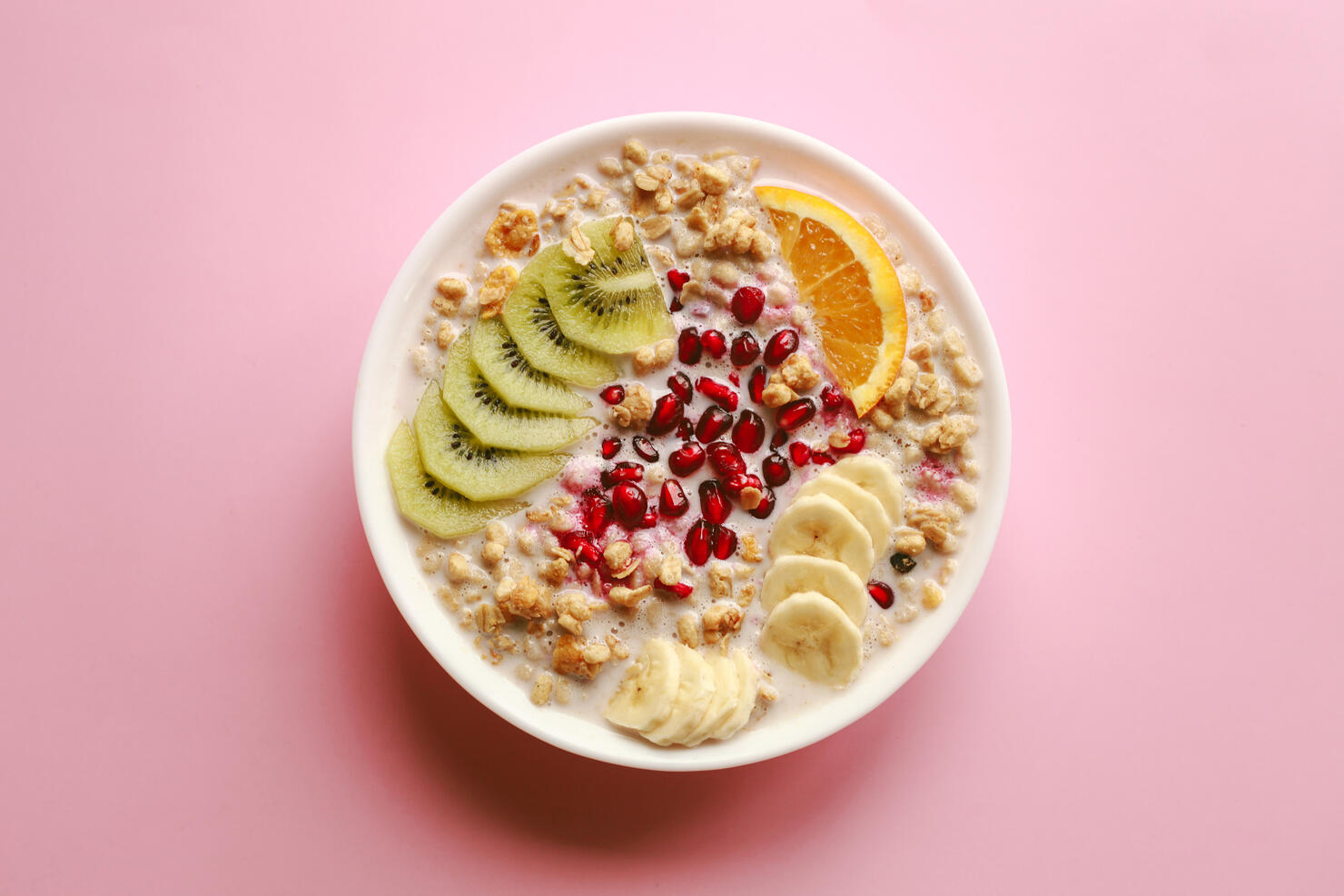 Breakfast cereal meal granola with milk, pomegranate, kiwi and orange in bowl and spoon over pink background