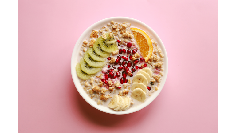 Breakfast cereal meal granola with milk, pomegranate, kiwi and orange in bowl and spoon over pink background