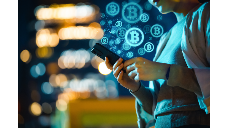 Mid-section of young Asian woman using smartphone in city at night, against illuminated street lights bokeh, working with Bitcoin technologies, investing or trading Bitcoin on cryptocurrency. Business on the go