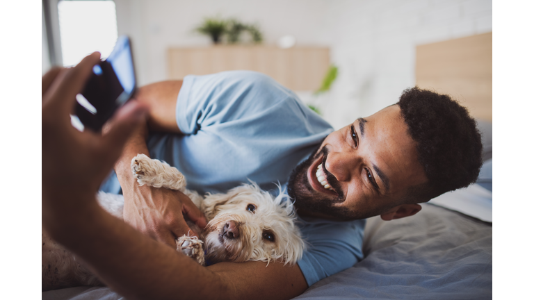 Portrait of young man with dog on bed indoors at home, taking selfie.