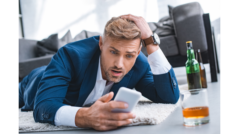 shocked businessman looking at smartphone and lying on carpet near glass with alcohol drink