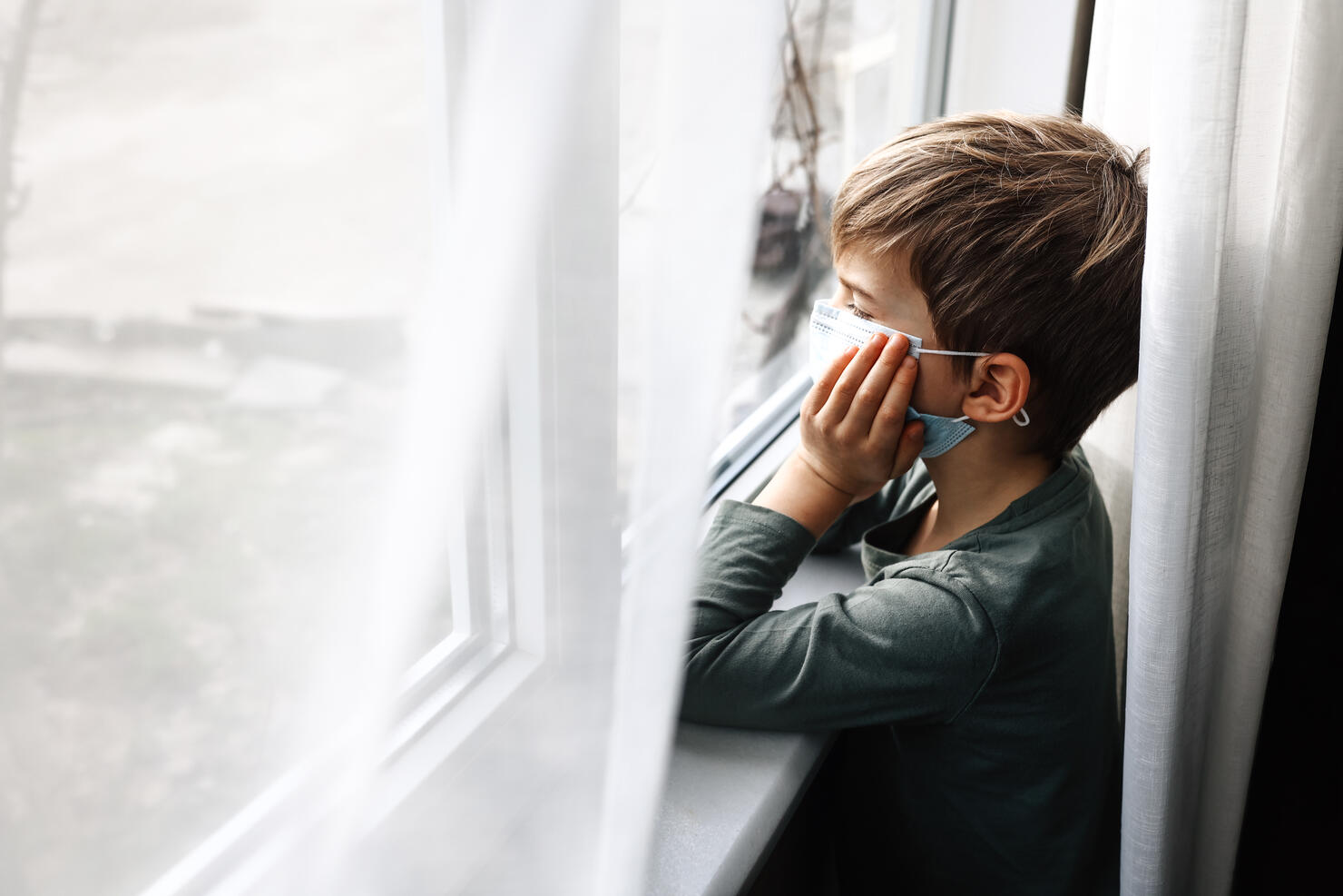 Small sad boy looking through the window during Coronavirus isolation.