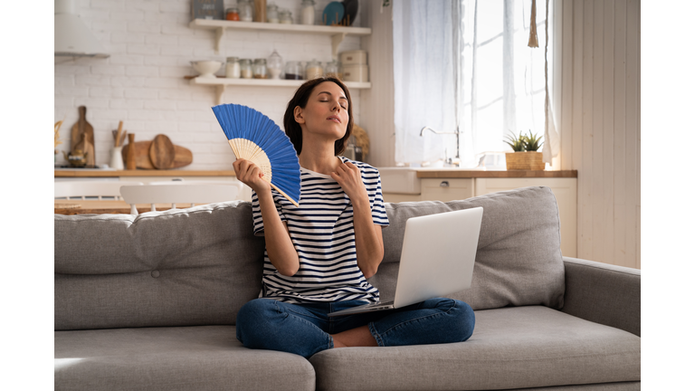 Young woman suffers from heatstroke flat without air-conditioner waving fan sitting on couch at home