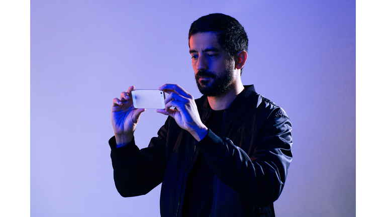 Man wearing jacket and holding cell phone illuminated by blue and red lights. Concept of man filming the police on his smartphone