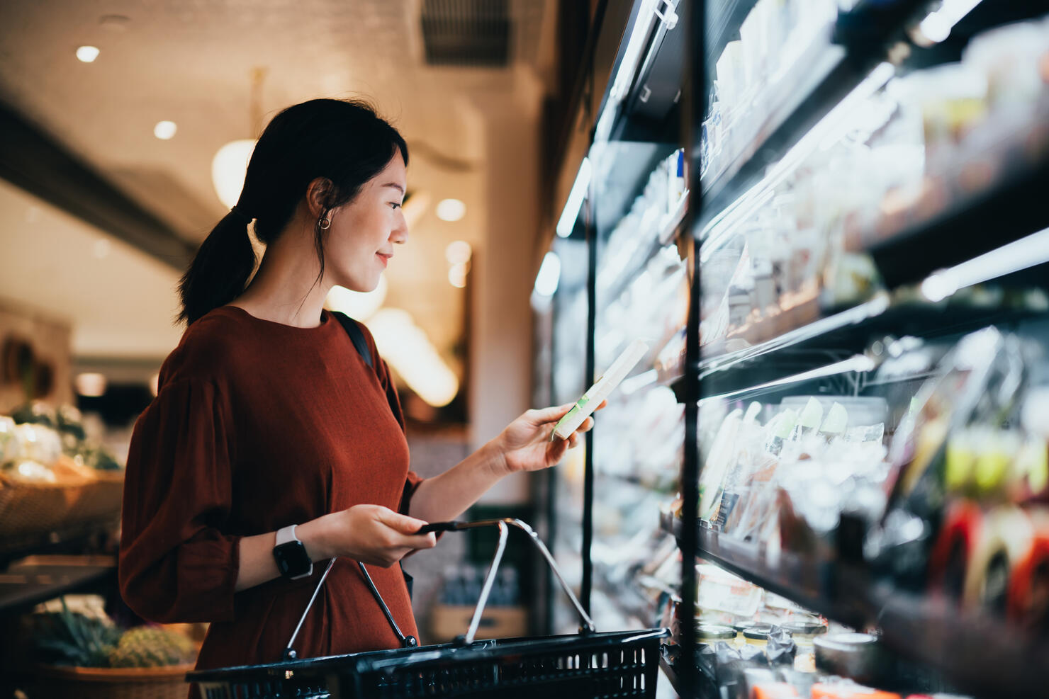 Young Asian woman carrying a shopping basket while grocery shopping in supermarket, standing along the dairy aisle, reading the nutrition label on a packet of fresh organic cheese. Making healthier food choices