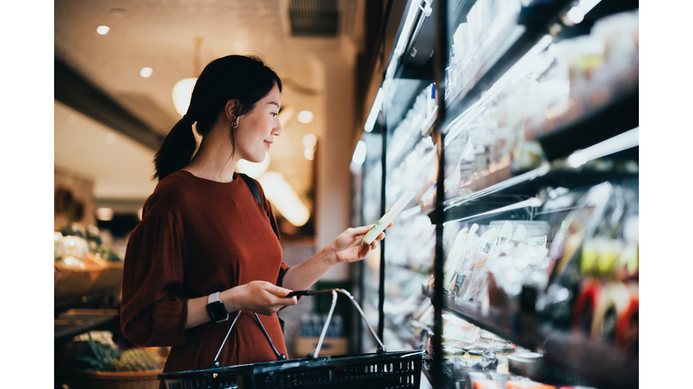 Young Asian woman carrying a shopping basket while grocery shopping in supermarket, standing along the dairy aisle, reading the nutrition label on a packet of fresh organic cheese. Making healthier food choices