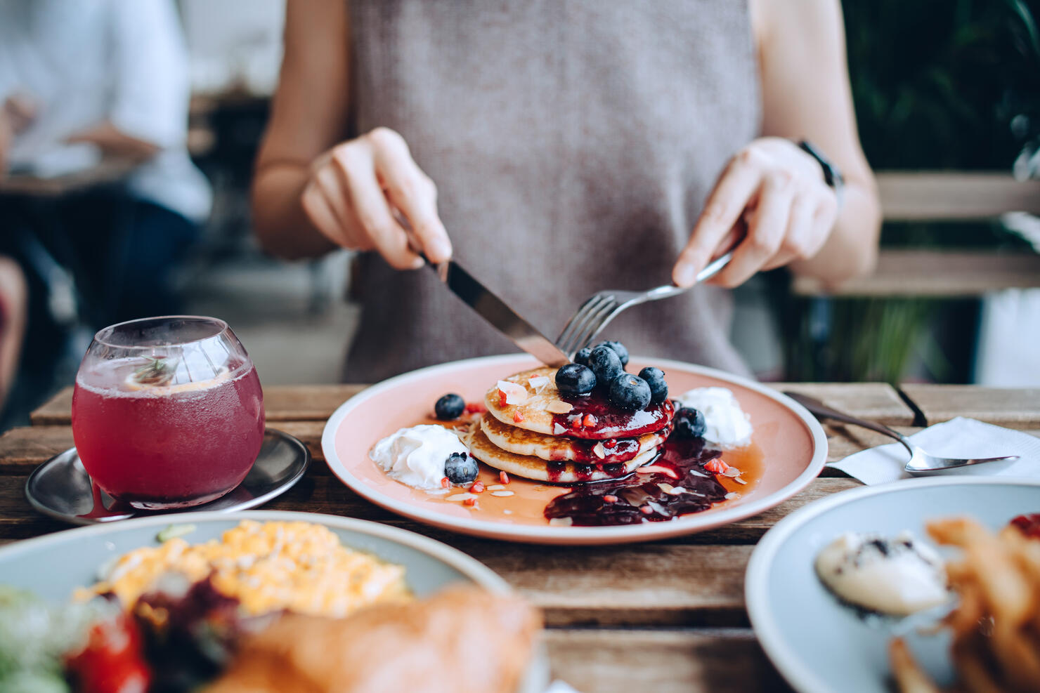 Close up of young woman sitting at dining table eating pancakes with blueberries and whipped cream in cafe, with English breakfast and french fries served on the dining table. Eating out lifestyle