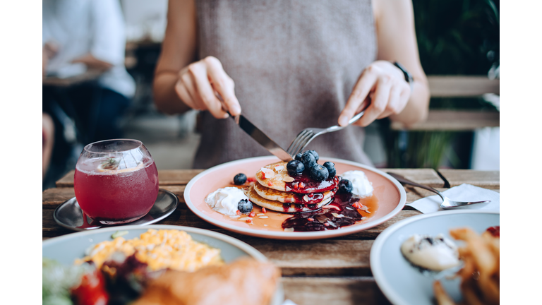 Close up of young woman sitting at dining table eating pancakes with blueberries and whipped cream in cafe, with English breakfast and french fries served on the dining table. Eating out lifestyle