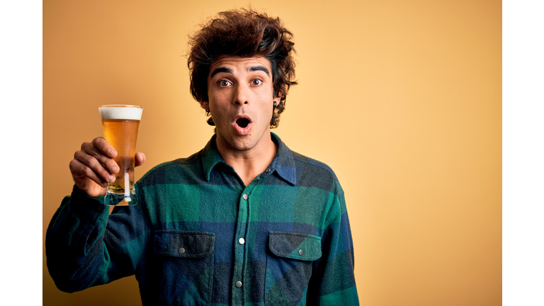 Young handsome man drinking glass of beer standing over isolated yellow background scared in shock with a surprise face, afraid and excited with fear expression