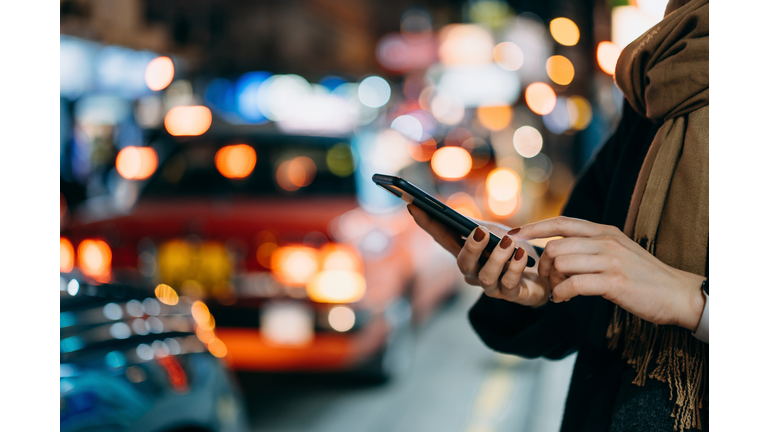 Close up of young woman using mobile app device on smartphone to arrange taxi ride in downtown city street, with illuminated busy city traffic scene during rush hour with traffic congestion in the evening