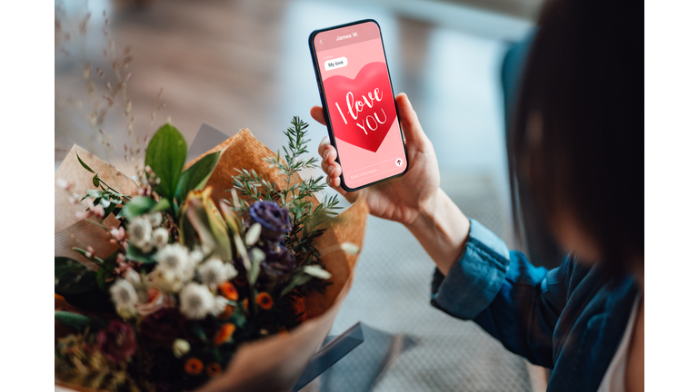 Over-the-shoulder view of a unrecognisable woman receiving flower bouquet and reading "I love you" message on mobile phone