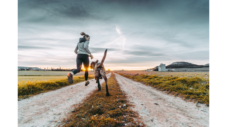 Woman and her dog running towards the sunset on a country road