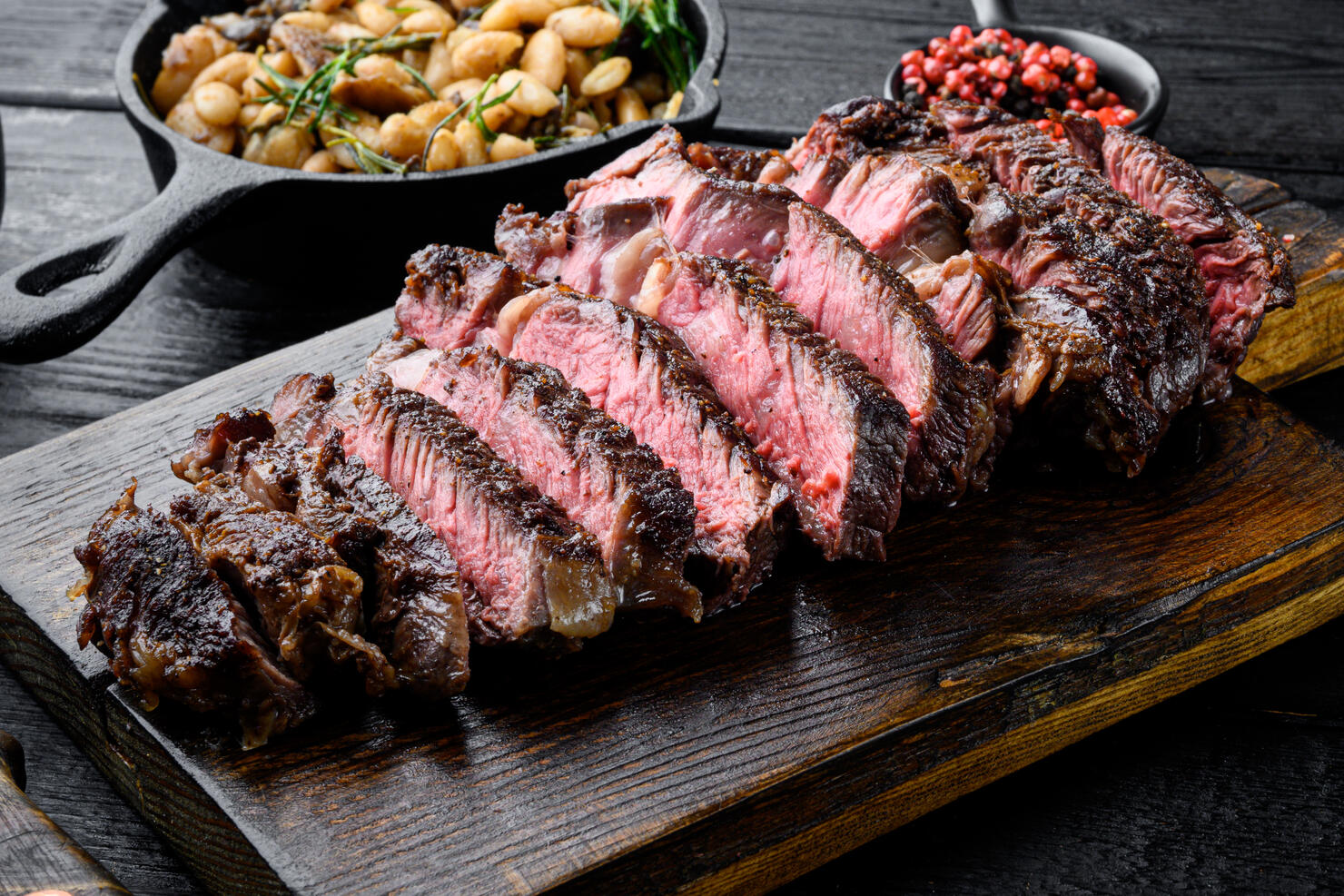 Sliced grilled meat steak Rib eye medium rare, on wooden serving board, with white beans and rosemary in cast iron pan, on black wooden table background