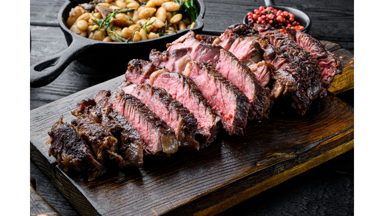 Sliced grilled meat steak Rib eye medium rare, on wooden serving board, with white beans and rosemary in cast iron pan, on black wooden table background