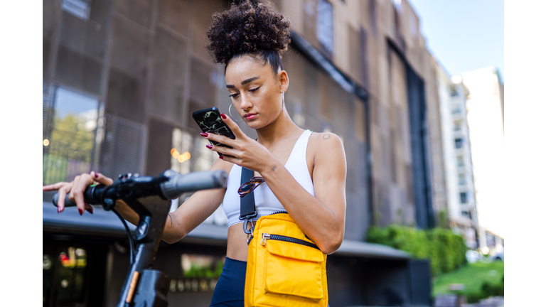 Mixed race young girl standing on the street with electric scooter while using smart phone