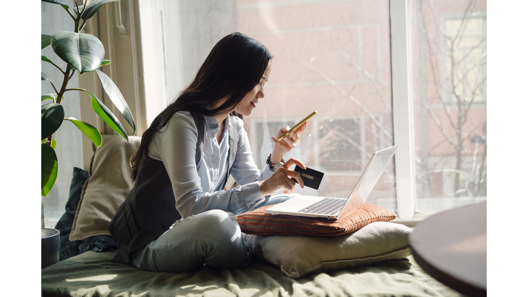 A middle-aged asian woman in blue jeans sitting on the bed in a yoga pose in front of a laptop