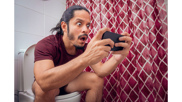 Young latin man smiling on the toilet using a smart phone at the bathroom