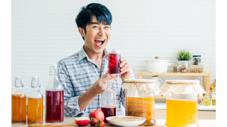 Portrait handsome man smiling with fresh and happiness, holding glass of Kombucha healthy fermented probiotic tea drinks with ingredients red apples, strawberries, mushroom scoby in kitchen at home.