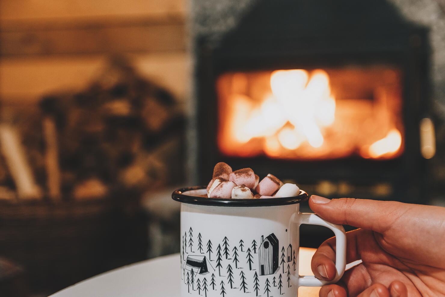 Woman's Hand Holding Cup Of Hot Drink In Front Of Fireplace In Cozy Cabin. Cocoa, Marshmallows.