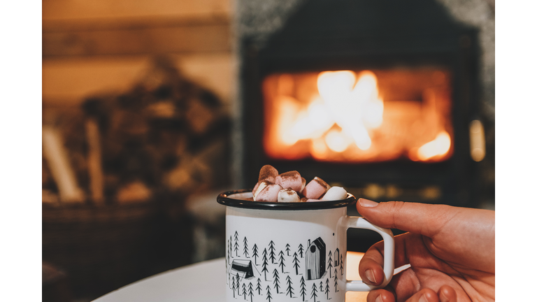 Woman's Hand Holding Cup Of Hot Drink In Front Of Fireplace In Cozy Cabin. Cocoa, Marshmallows.